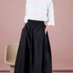 PAPILLONE pleated midi skirt sewing pattern: black high-waisted skirt with broad box pleats at the sides, model in a loose white blouse and light heeled sandals against a grey studio backdrop.