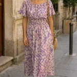Patrón de costura del vestido DUNE: vestido midi lila con flores, mangas cortas abullonadas y cintura fruncida, en una calle.