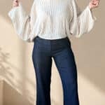 Patrón de costura pantalón acampanado AUDE: mujer sonriente con los brazos en alto, jersey blanco de mangas anchas
