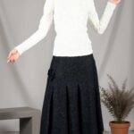 LANGE flared midi skirt sewing pattern: studio front view of a model in a black speckled skirt with soft vertical pleats and fabric flower, white top and lace-up boots.