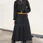 LANGE pleated skirt sewing pattern: outdoor portrait of a woman in a charcoal midi skirt with wide front pleats, black blouse, mustard-yellow belt and tall black boots.