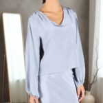 PATY V-neck blouse sewing pattern: light powder-blue flowing top with gentle gathers near the neckline, long sleeves and narrow cuffs, worn with a matching skirt against a gray studio wall.