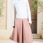 AVA box-pleat midi skirt sewing pattern: model wearing a peach-pink patterned flared skirt with a central front pleat, white textured top and pale knee-high boots.